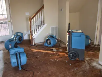 Blue air movers and a dehumidifier drying a stripped floor beside a staircase in a Garner area home